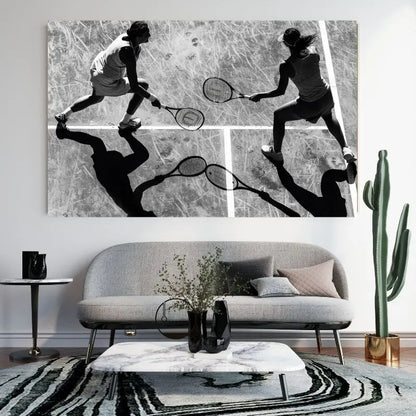 Un Tableau Cours Tennis Doublette Noir et Blanc est exposé au-dessus d'un canapé gris moderne, avec un cactus à droite et une table basse blanche devant, encapsulant une décoration intérieure chic et de l'art sportif.