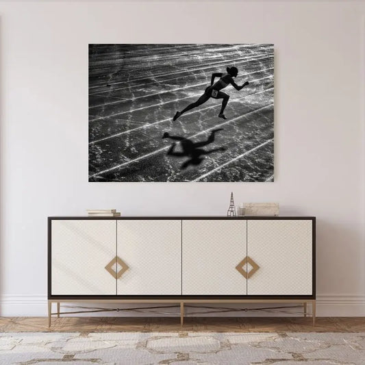 Une photographie en noir et blanc accrochée au mur montre un athlète en pleine course, projetant une ombre sur la piste. Ce Tableau Athlétisme Coureur Noir et Blanc est exposé au-dessus d'une console moderne aux poignées géométriques.