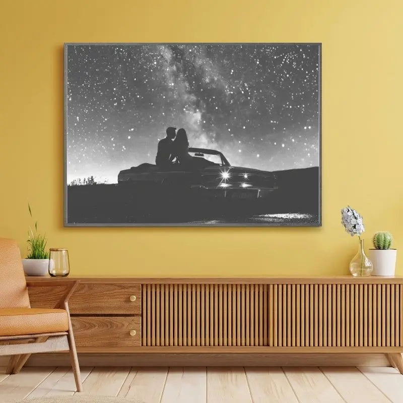 Le Tableau Couple Amoureux Voiture Ciel Etoilé Noir et Blanc, représentant un couple assis sur le capot d'une voiture sous un ciel étoilé, orne le mur jaune au-dessus du buffet en bois au décor minimaliste, créant une atmosphère romantique.