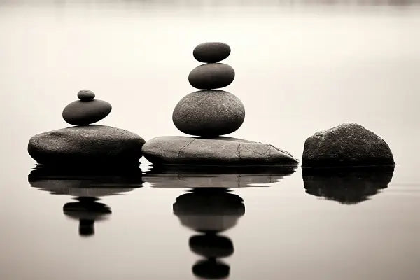 Trois piles de pierres équilibrées reposent sur de gros rochers au bord de l'eau, créant un Tableau Zen Minimaliste Pierre Pile Cairns Relaxation Noir et Blanc. Leurs reflets sont visibles dans l'eau calme, ajoutant à l'ambiance de détente et d'équilibre.