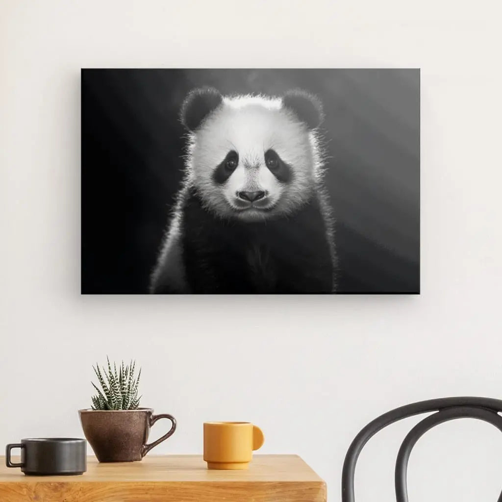 Une superbe photographie Noir et Blanc d'un panda est accrochée au mur au-dessus d'une table en bois ornée d'une petite plante, d'une tasse marron et d'une tasse jaune. Une chaise noire est partiellement visible à droite, ajoutant à ce charmant Tableau Panda Portrait Jungle Animal Noir et Blanc.