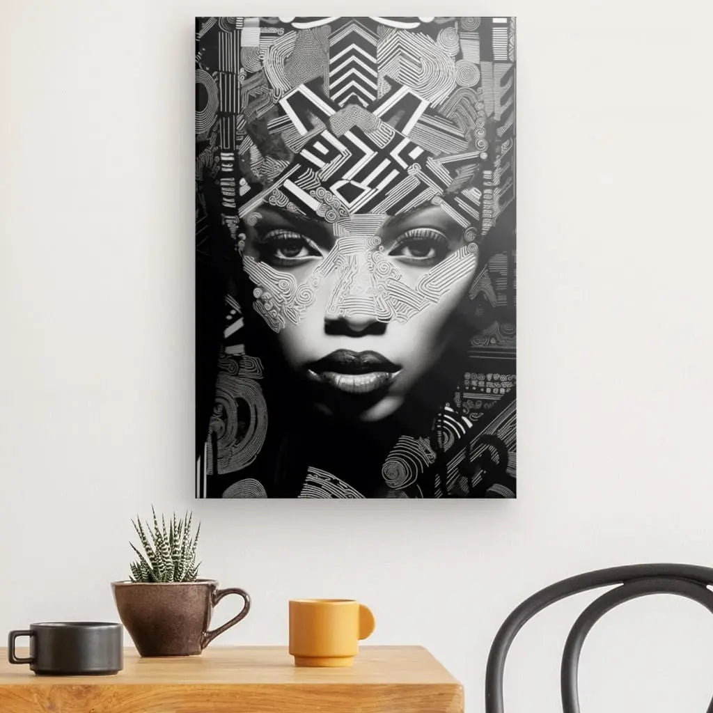 Un Tableau Portrait Femme Africaine Art Ethnique Noir et Blanc en noir et blanc représentant un visage de femme avec des motifs géométriques est accroché au mur au-dessus d'une table en bois avec une petite plante et deux tasses. Une chaise est partiellement visible à droite, accentuant le portrait captivant.