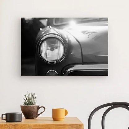 Une photo en noir et blanc du phare et de l'aile avant d'un Tableau Voiture Ancienne Phare Rond Vintage Design Noir et Blanc est accrochée sur un mur blanc. En dessous, une table contient une plante et deux tasses. Vue partielle d'une chaise à droite.