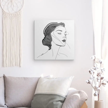 Un portrait de femme minimaliste en dessin au trait est accroché sur un mur blanc au-dessus d'un canapé gris avec un oreiller blanc. Une tenture murale en macramé se trouve à gauche et une plante blanche à droite, complétant le Tableau Portrait Femme Diva Dessin Moderne Visage Traits Noir et Blanc.