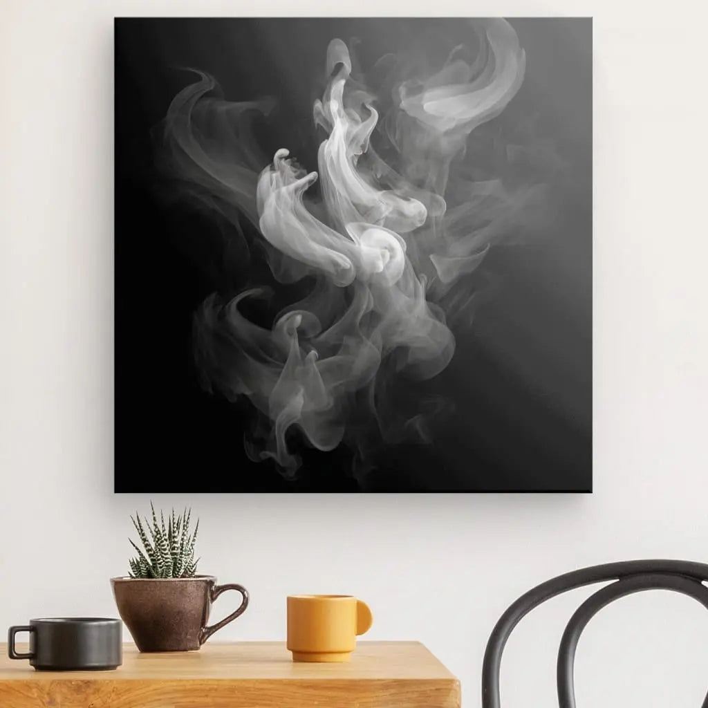 Une œuvre d'art abstraite en noir et blanc, Tableau Fumée Blanche Fond Noir Design Moderne Vapeurs et Effluves représentant de la fumée tourbillonnante, est suspendue sur un mur blanc au-dessus d'une table en bois avec une petite plante, des tasses marron et jaune et le dossier d'une chaise visible.