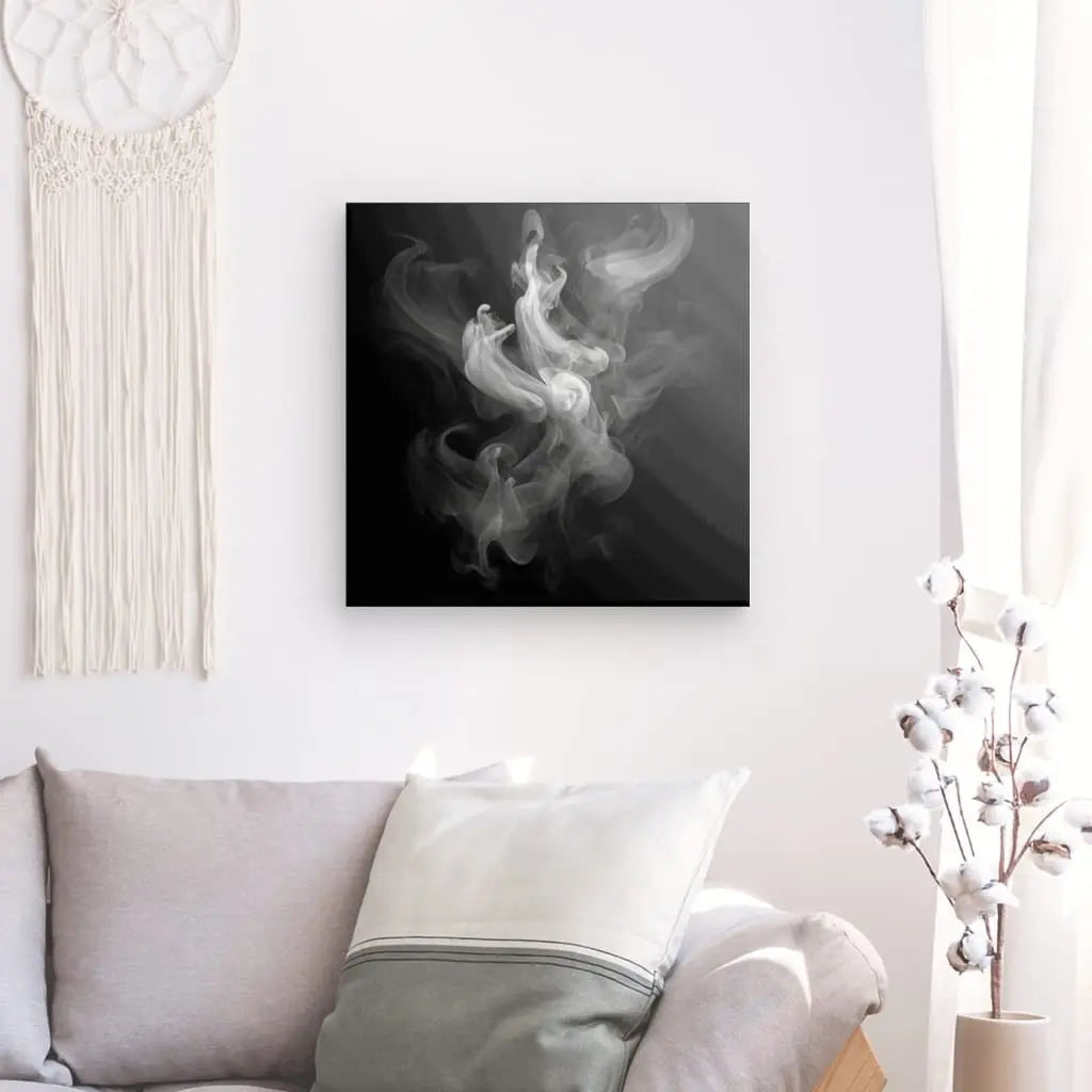 Un Tableau Fumée Blanche Fond Noir Design Moderne Vapeurs et Effluves encadré, incarnant une esthétique de tableau noir et blanc, est accroché sur un mur blanc au-dessus d'un canapé gris clair avec un oreiller blanc et un oreiller à rayures crème. Une tenture murale en macramé et une plante en pot ajoutent au design moderne.