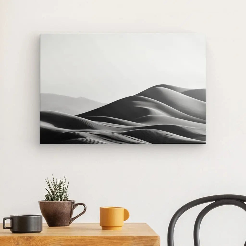 Un Tableau Photographique Dunes de Sable Noir et Blanc est accroché sur un mur blanc au-dessus d'une table en bois contenant un petit cactus, une tasse marron et une tasse jaune. Une chaise noire est partiellement visible à droite.