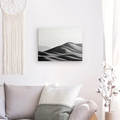 Un salon avec un canapé beige, un coussin, une décoration murale suspendue en macramé et un Tableau Photographique Dunes de Sable Noir et Blanc capture l'essence de l'élégance minimaliste. Un vase blanc avec des branches de coton complète le décor serein.