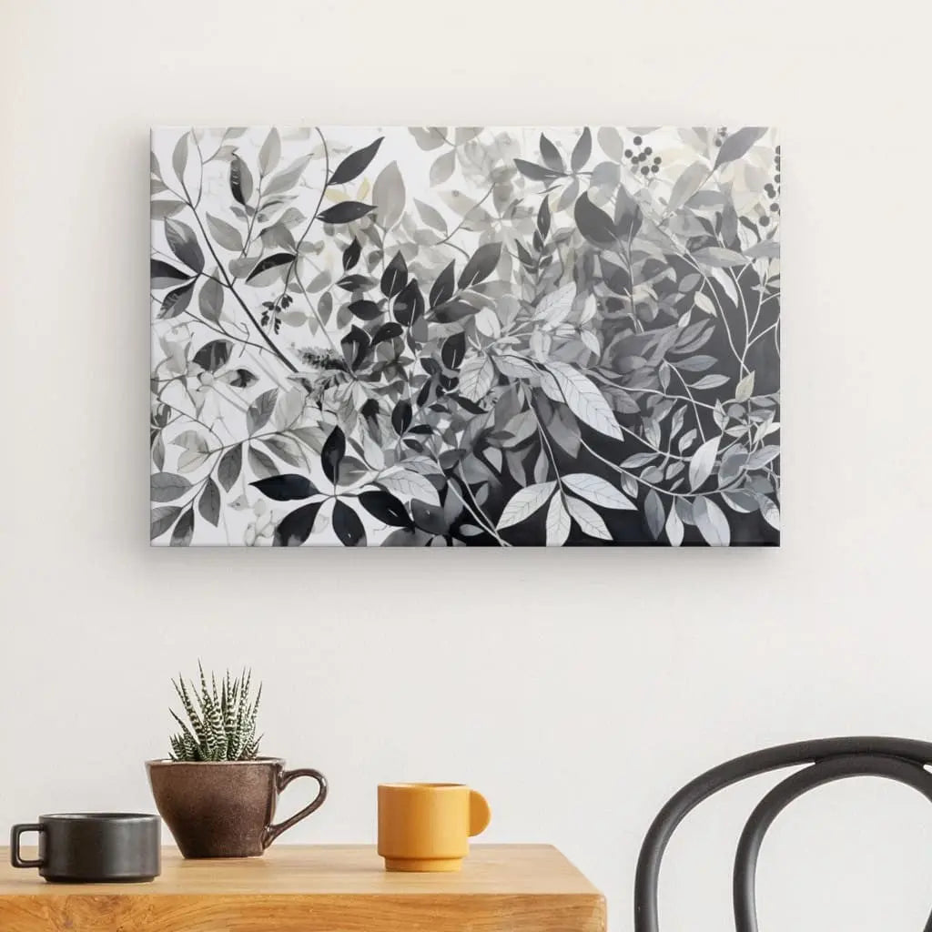 Un Tableau Plantes Feuilles Végétaux Nature Noir et Blanc mural au-dessus d'une table en bois avec une petite plante en pot, une tasse à café marron et une tasse jaune. Une chaise noire est partiellement visible sur la droite, rehaussant la décoration intérieure sophistiquée.