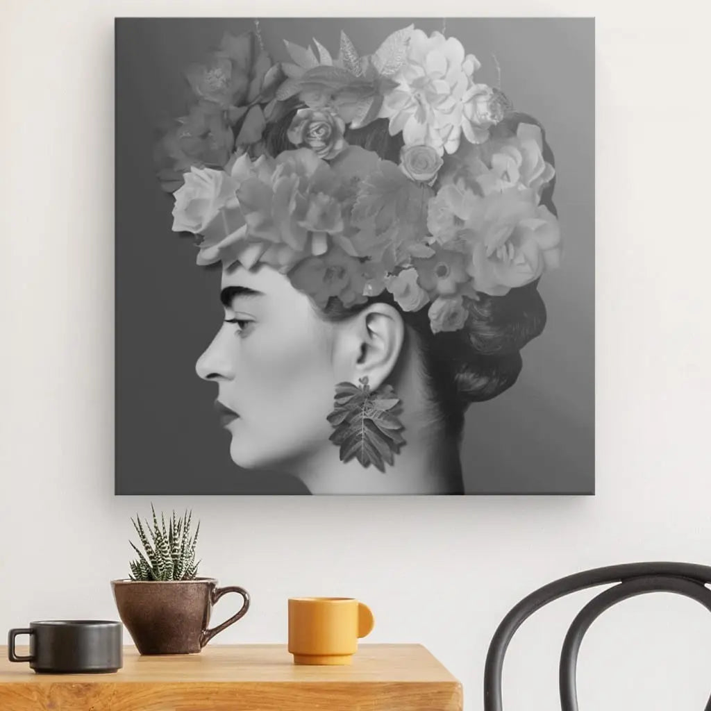 Un saisissant Tableau Profil Frida Fleurs Cheveux Rose Pivoine Portrait Noir et Blanc, cette photographie en noir et blanc d'une femme de profil, ornée d'une coiffe fleurie et de grandes boucles d'oreilles fleuries, est suspendue au-dessus d'une petite table avec deux tasses et une plante en pot. Cette œuvre artistique dégage une élégance intemporelle.