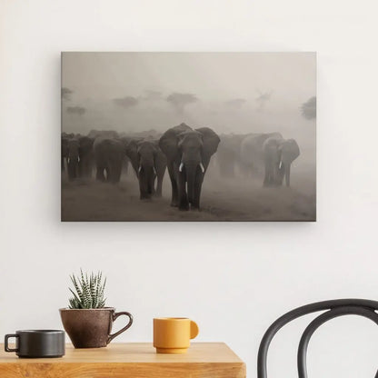 Un Tableau Troupeau Famille Elephants Savane Noir et Blanc est accroché sur un mur blanc au-dessus d'une petite table en bois avec une plante et deux tasses.