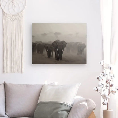 Un salon comprend un canapé avec des coussins gris clair, une tenture murale et une photo encadrée du Tableau Troupeau Famille Elephants Savane Noir et Blanc marchant dans un paysage poussiéreux. Un décor végétal en acrylique se trouve dans le coin de la pièce, complétant l’esthétique globale.