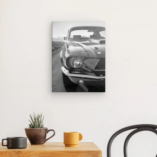 Une photo en noir et blanc d'une voiture classique, peut-être une Mustang Tableau Voiture Vintage Noir et Blanc Muscle Car, est montée sur un mur blanc au-dessus d'une table en bois avec une plante en pot et deux tasses. Une chaise noire est partiellement visible à droite, créant une élégante scène de tableau noir et blanc.