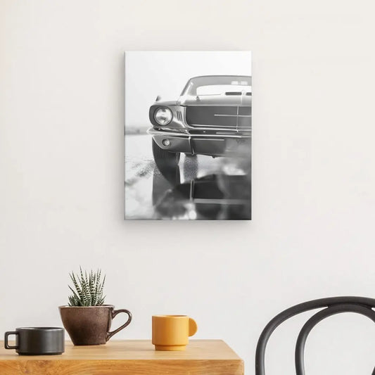 Un Tableau Voiture Ancienne Noir et Blanc Mustang est accroché sur un mur blanc au-dessus d'une table en bois. Sur la table se trouvent une petite plante en pot, une tasse marron et une tasse jaune. Une chaise noire est partiellement visible.