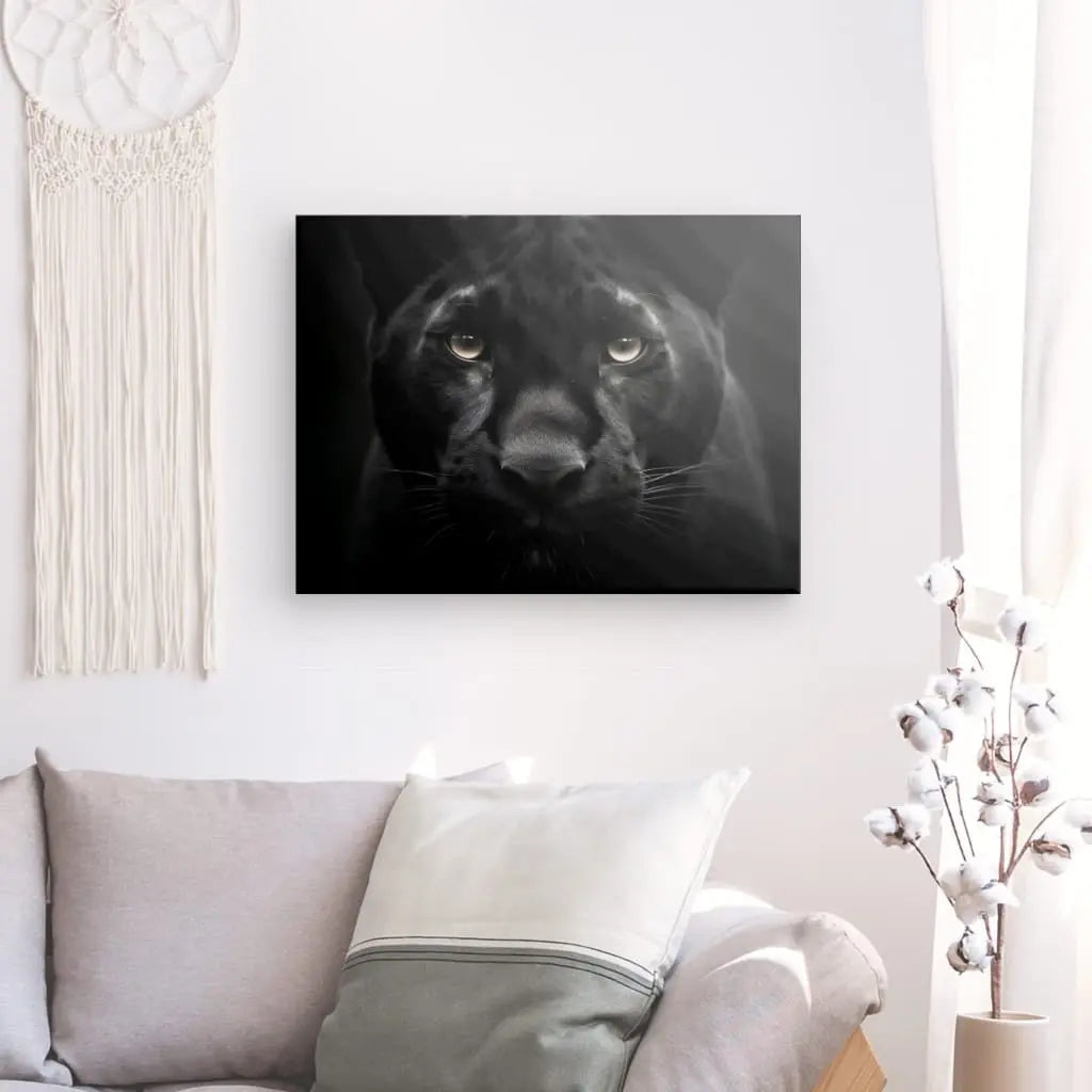 Un *Tableau Panthère Noire Regard Percant Portrait Animal Sauvage Noir et Blanc* est exposé au-dessus d'un canapé de couleur claire dans une pièce baignée de lumière naturelle et dotée d'une tenture en macramé.