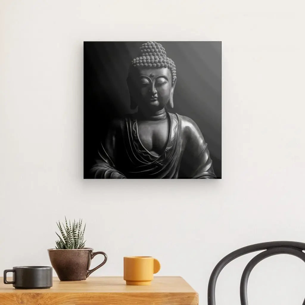 Photographie en noir et blanc d'une statue de Bouddha sur une toile carrée, créant le Tableau Zen Bouddha Méditation Noir et Blanc. Il est accroché à un mur blanc au-dessus d'une table en bois clair avec une petite plante, une tasse marron et une tasse jaune à côté d'une chaise noire. Cette subtile décoration intérieure apporte de la quiétude à l'espace.