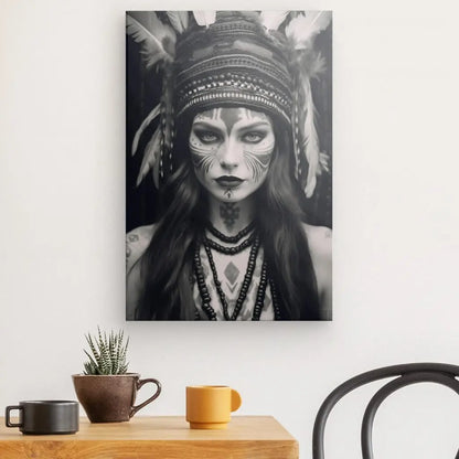 Un superbe portrait en noir et blanc d'un Tableau Femme Indienne Portrait Noir et Blanc avec une peinture faciale d'inspiration tribale et une coiffe à plumes est accroché sur un mur blanc. Cette œuvre d'art saisissante est magnifiquement exposée au-dessus d'une table en bois ornée d'une plante en pot et de tasses, tandis qu'une chaise jette un coup d'œil dans le cadre.