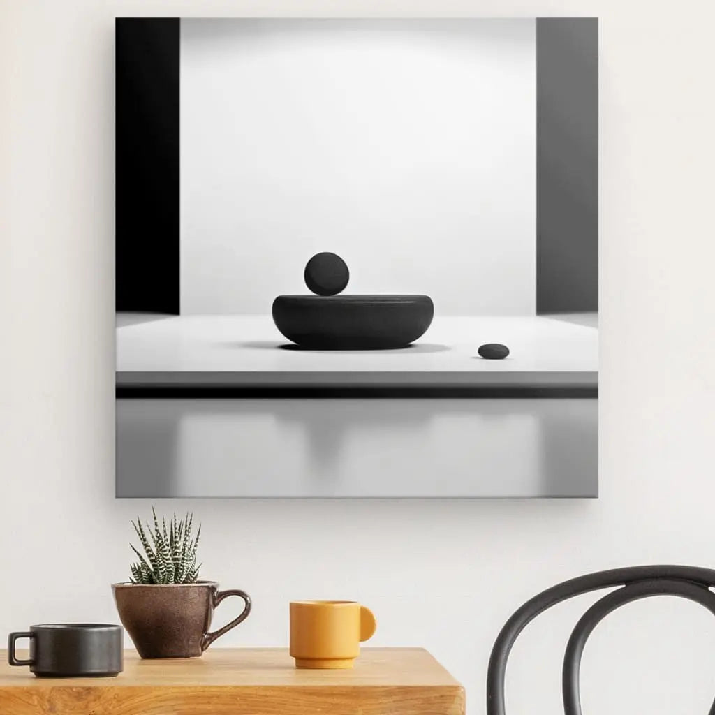 Une photographie minimaliste en noir et blanc capture un objet rond dans un bol, avec un autre petit objet rond à côté. Le Tableau Carré Relaxation et Paix Noir et Blanc s'affiche sur un mur blanc au-dessus d'une table en bois ornée de tasses et d'une plante, évoquant la détente.