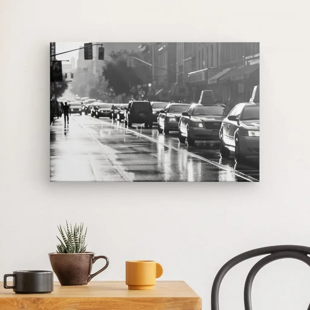 Photo en noir et blanc d'une rue de ville au trottoir mouillé, bordée de voitures pour la plupart à l'arrêt et quelques piétons au loin, rappelant un Tableau Rue New-Yorkaise Taxis Wall Street Noir et Blanc, posé sur un mur blanc au dessus d'une petite table en bois avec une plante et deux mugs.