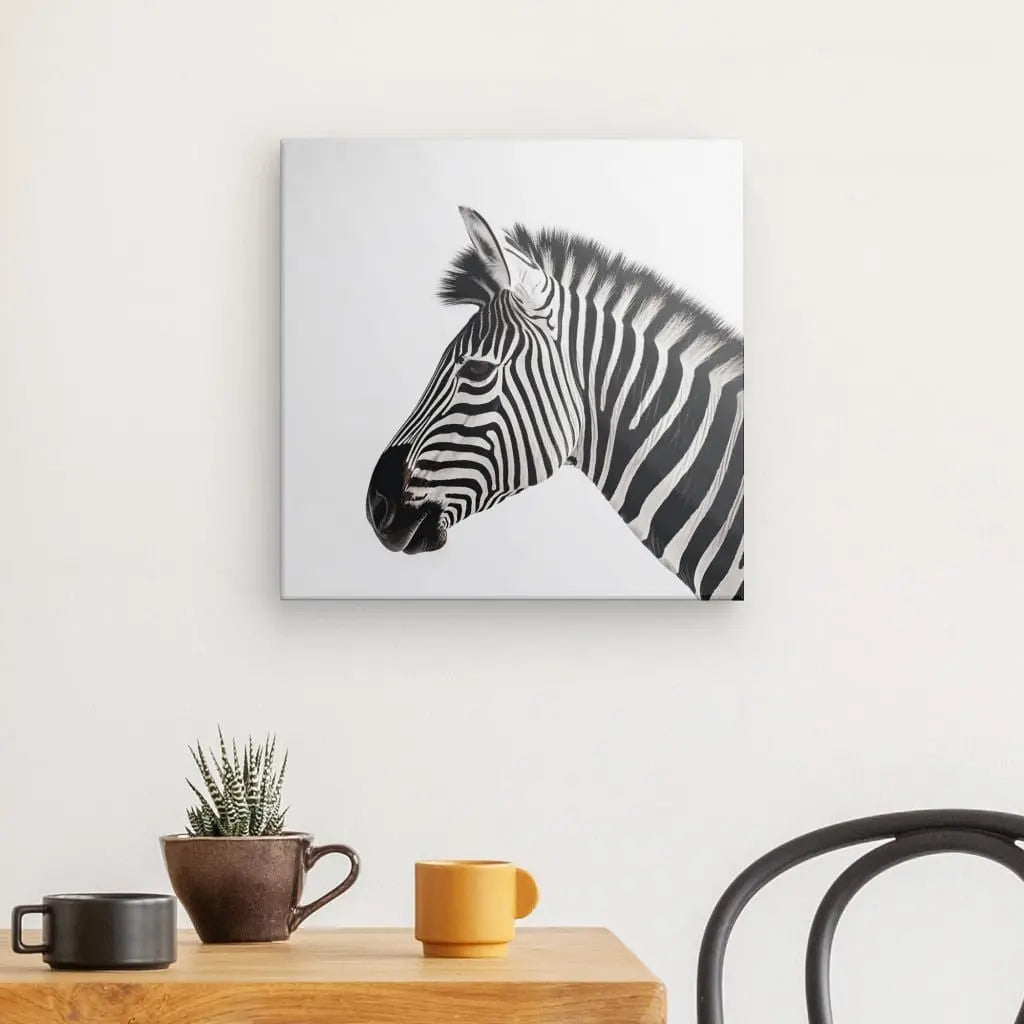 Un Tableau Carré Profil Zèbre Savane Noir et Blanc est accroché sur un mur blanc au-dessus d'une table en bois avec une petite plante en pot et deux tasses. Le bord d'une chaise noire est visible à droite, rehaussant la décoration intérieure chic.