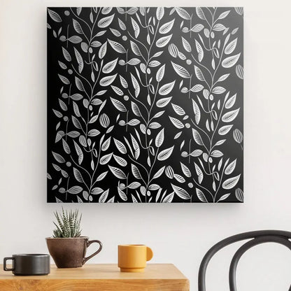 Une décoration murale monochrome présentant des motifs de feuilles blanches sur fond noir incarne l’esthétique noir et blanc. Ci-dessous, il y a une table en bois avec une plante en pot, une tasse marron et une tasse jaune. Une chaise en métal noir est visible à droite, renforçant l'ambiance Tableau Végétation Plantes Grimpantes Minimalistes Noir et Blanc.