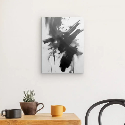 Un Tableau Abstrait Asiatique Traces Peinture Noires en noir et blanc est accroché sur un mur blanc au-dessus d'une table en bois. La table, faisant écho à une ambiance art zen, comporte une petite plante en pot et deux tasses, une marron et une jaune. Une chaise noire est partiellement visible.