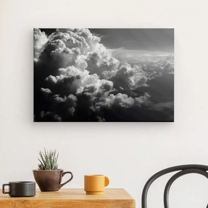 Une photographie en noir et blanc d'un ciel nuageux, créant un Tableau Cloudy Nuages Coton Noir et Blanc, est accrochée sur un mur blanc au-dessus d'une table en bois avec une plante en pot, une tasse marron et une tasse jaune. Une chaise noire est partiellement visible à droite, ajoutant à la décoration intérieure monochrome noir et blanc.