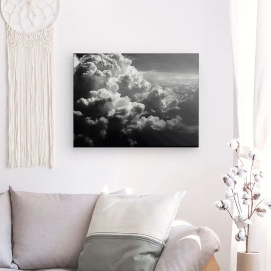 Un espace de vie avec un canapé gris, des coussins et une décoration en cotonnier. Un Tableau Cloudy Nuages Coton Noir et Blanc est accroché sur le mur blanc à proximité d'une tenture murale en macramé.