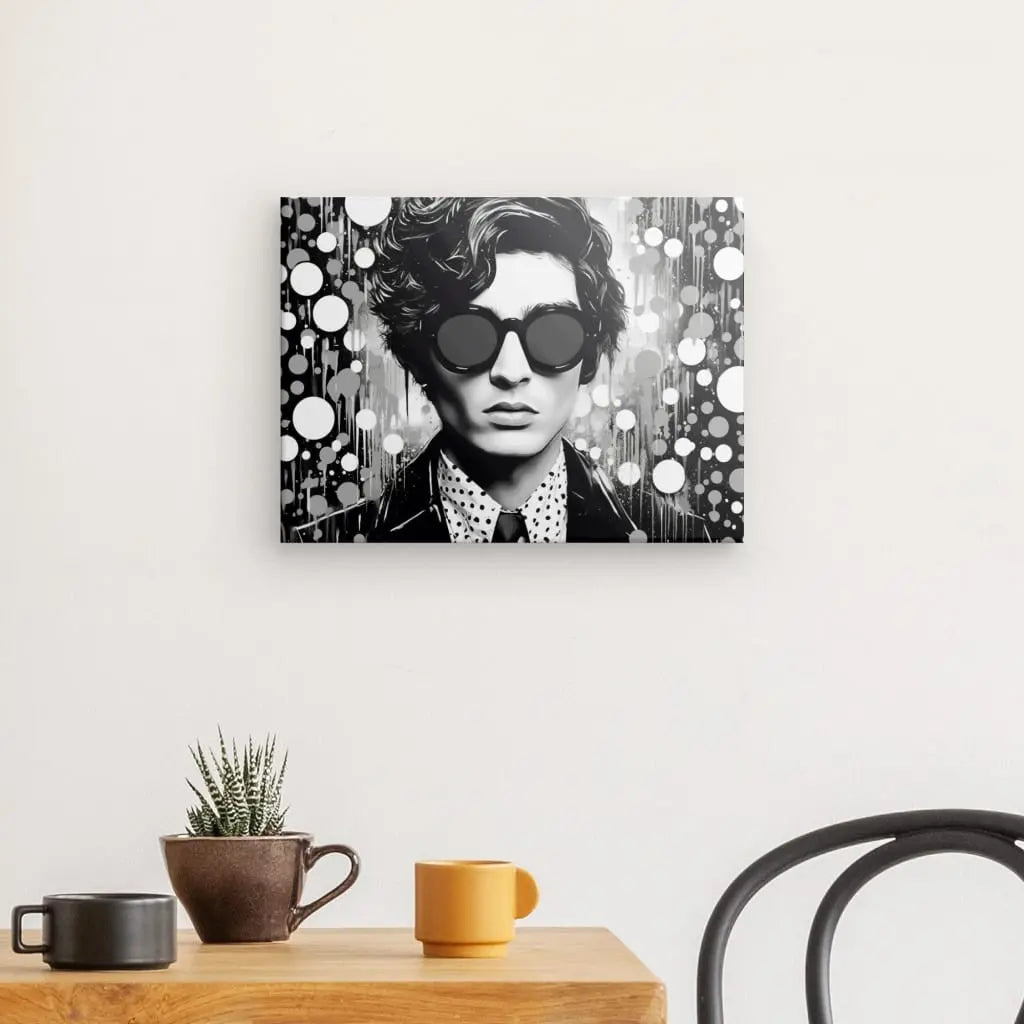 Un portrait en noir et blanc d'une personne portant des lunettes de soleil sombres, rendu dans un style esthétique pop art, est accroché sur un mur blanc au-dessus d'une table en bois avec des plantes en pot et deux tasses. Une vue partielle d'une chaise noire est visible. L'œuvre exposée est le "Tableau Homme Pop Art Bulles Noir et Blanc.