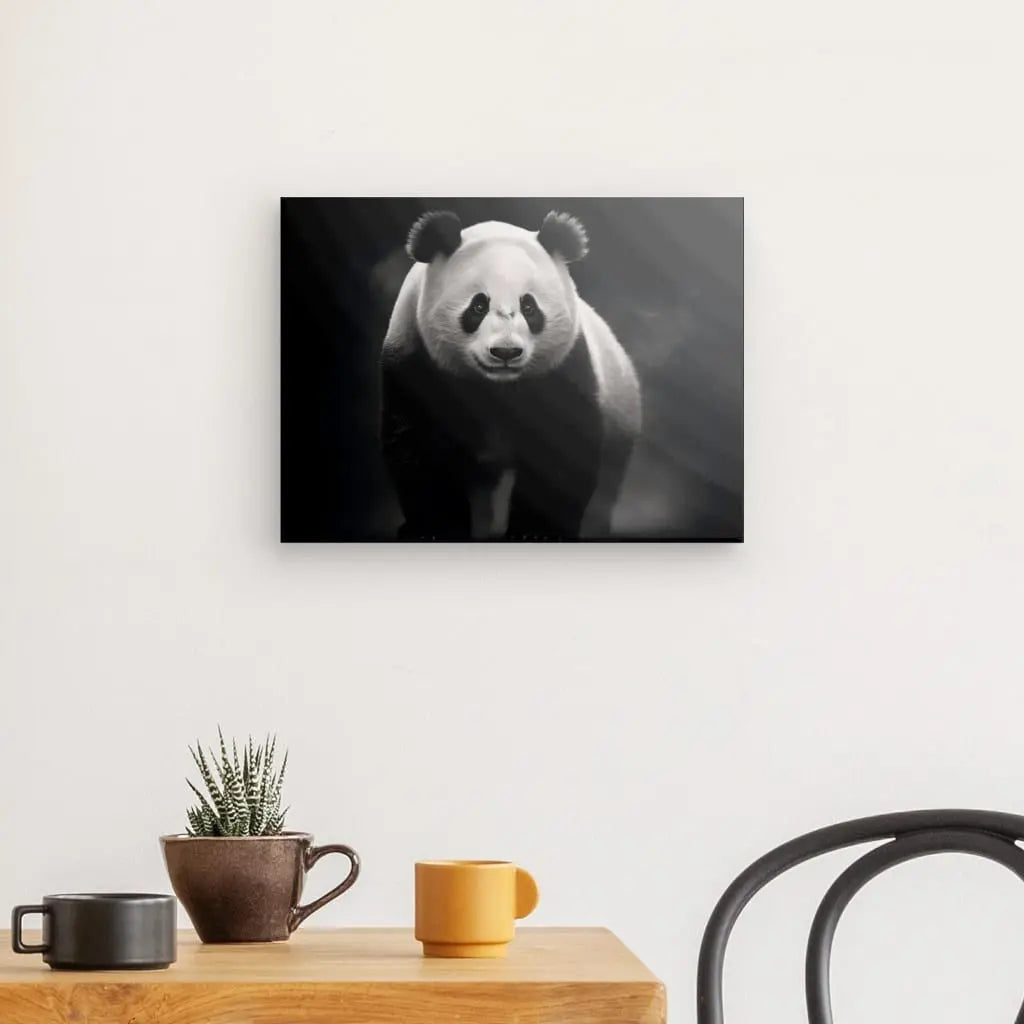 Une photo en noir et blanc d'un Tableau Panda Sauvage Animal Noir et Blanc est accrochée sur un mur blanc au-dessus d'une table en bois. Sur la table se trouvent un cactus en pot et deux tasses, une jaune et une marron. Une chaise noire est partiellement visible, créant une ambiance sereine et plus animale.