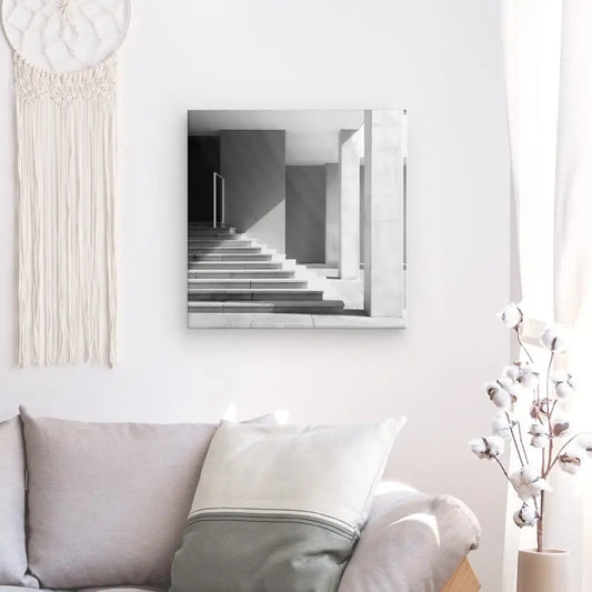 Un salon minimaliste comprend un canapé gris avec des oreillers blancs et gris, une tenture murale tissée et une photographie encadrée Tableau architecture Bâtiment Pierre Moderne Noir et Blanc sur un mur blanc à côté d'une fenêtre.