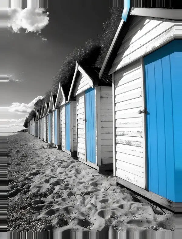 Rangée de cabanes de plage blanches avec portes bleu vif sur un rivage sablonneux sous un ciel partiellement nuageux. L'image est principalement en noir et blanc, à l'exception des portes bleues, créant un effet d'art mural qui ressemble à Tableau Cabanes Plage Noir Blanc Bleu.