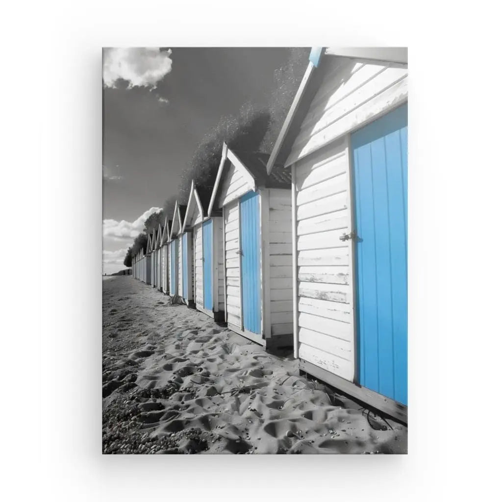 Rangée de cabines de plage aux murs blancs et portes bleues, posées sur une plage de sable et un ciel partiellement nuageux. Ce Tableau Cabanes Plage Noir Blanc Bleu est en grande partie en noir et blanc, à l'exception des portes bleues frappantes qui captivent le spectateur.