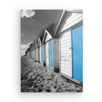 Rangée de cabines de plage aux murs blancs et portes bleues, posées sur une plage de sable et un ciel partiellement nuageux. Ce Tableau Cabanes Plage Noir Blanc Bleu est en grande partie en noir et blanc, à l'exception des portes bleues frappantes qui captivent le spectateur.