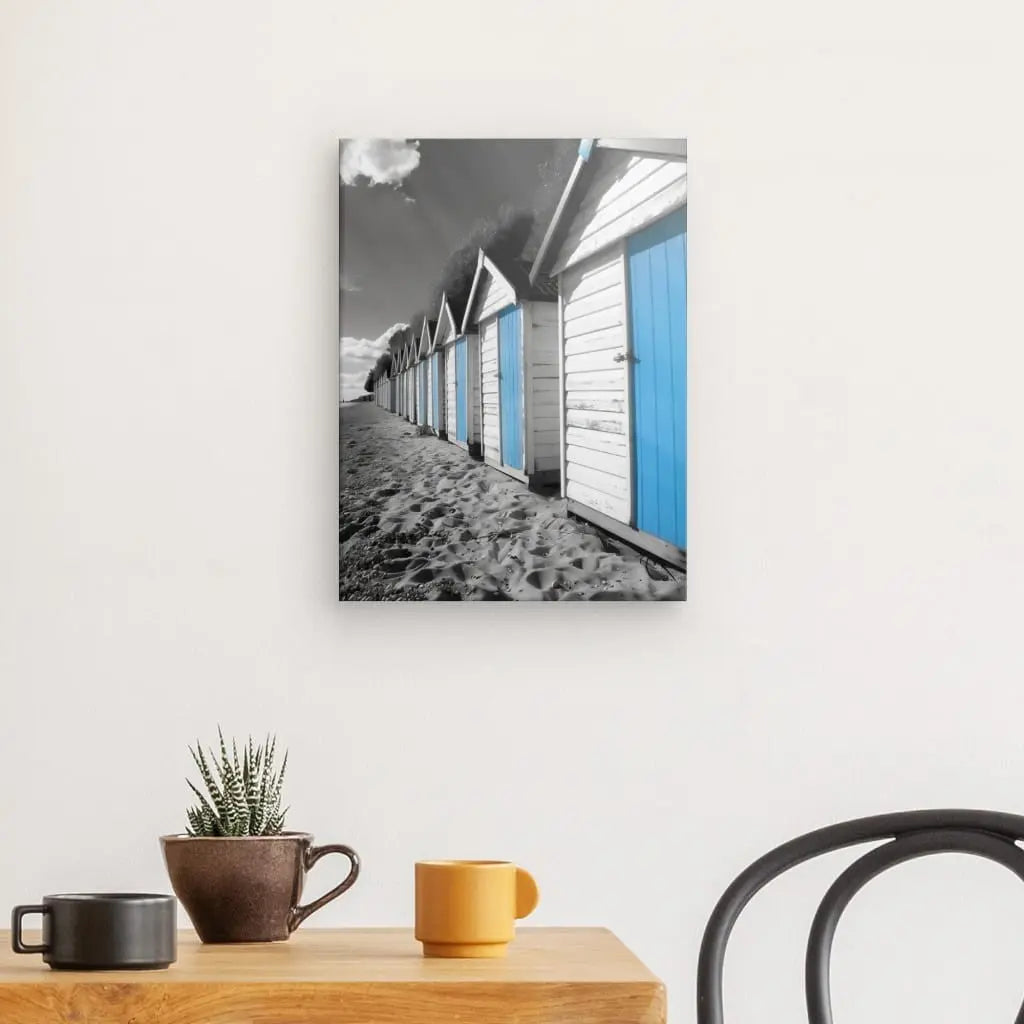 Un Tableau Cabanes Plage Noir Blanc Bleu est accroché sur un mur blanc au-dessus d'une table en bois ornée d'une petite plante, d'une tasse marron et d'une tasse orange. Une chaise noire est partiellement visible à droite.