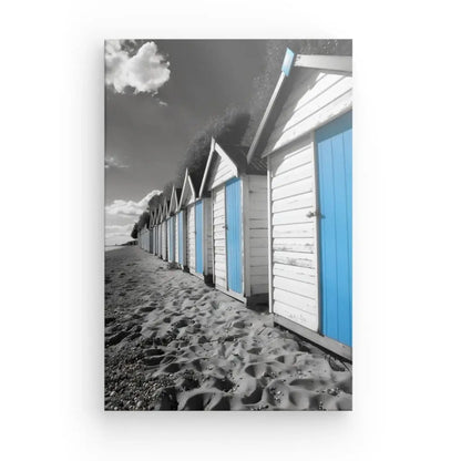 Une rangée de cabines de plage identiques aux portes bleues se dresse sous un ciel partiellement nuageux, créant un Tableau Cabanes Plage Noir Blanc Bleu. La vue s'étend vers l'horizon, avec du sable s'étendant au premier plan comme une peinture murale.