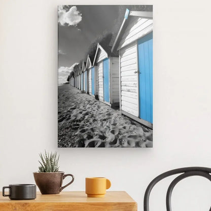 Une photographie d'une rangée de cabines de plage aux portes bleues, un charmant Tableau Cabanes Plage Noir Blanc Bleu, est accrochée à un mur blanc au-dessus d'une table contenant une plante, une tasse jaune et un bol marron.