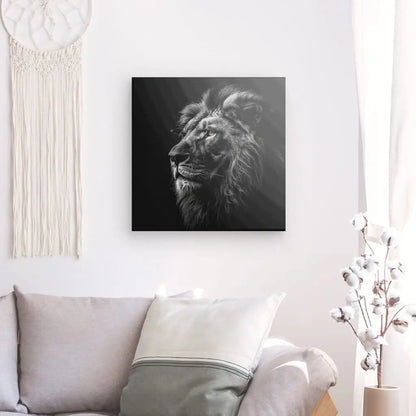 Une photographie en noir et blanc d'un lion, faisant partie de la collection Tableau Lion Roi Jungle Noir et Blanc Yeux Marrons, est accrochée sur un mur blanc au-dessus d'un canapé gris avec des oreillers blancs et une couverture légère drapée sur le dossier. Une tenture murale en macramé à gauche ajoute une touche de charme à cette élégante décoration d'intérieur.