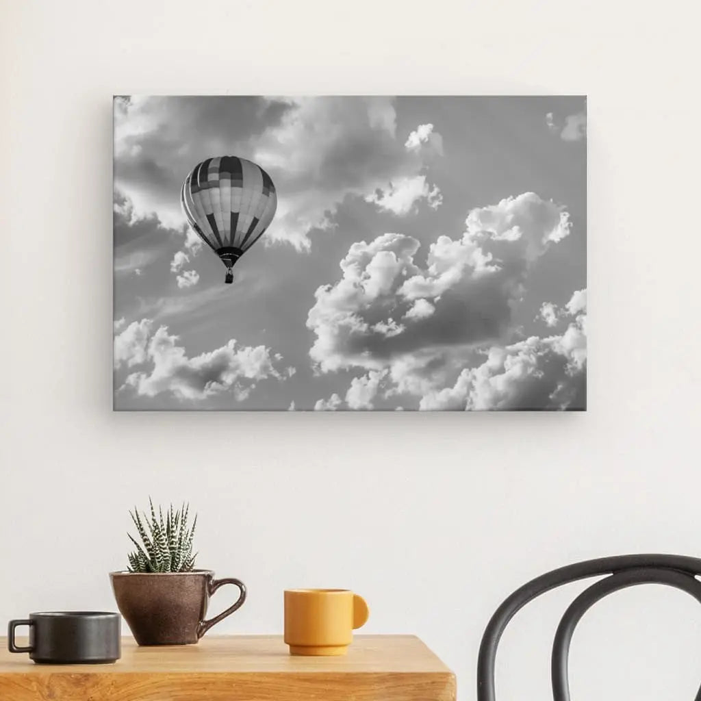 Un Tableau Montgolfière Aérostat Paysage Ciel Noir et Blanc représentant une montgolfière dans le ciel est accroché sur un mur blanc au-dessus d'une table en bois avec deux tasses, une plante en pot et une chaise noire.