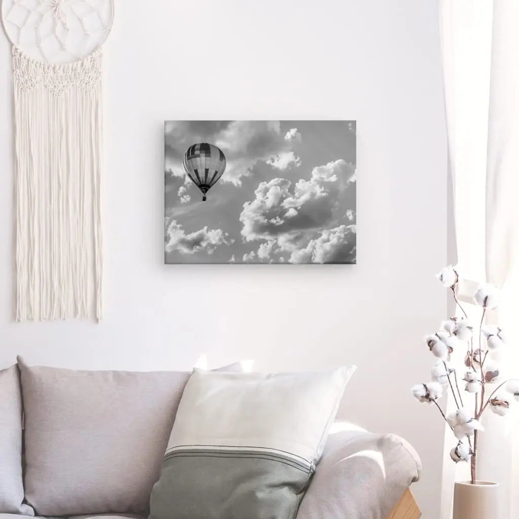 Un salon avec un canapé gris, un coussin blanc et un Tableau Montgolfière Aérostat Paysage Ciel Noir et Blanc en noir et blanc au mur. Un macramé décoratif est accroché à côté de la photographie.