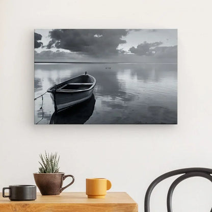 Un Tableau Barque Lac Paisible Noir et Blanc noir et blanc sur une eau calme sous un ciel nuageux, affiché sur un mur blanc au-dessus d'une table avec une plante en pot, une tasse et un bol.