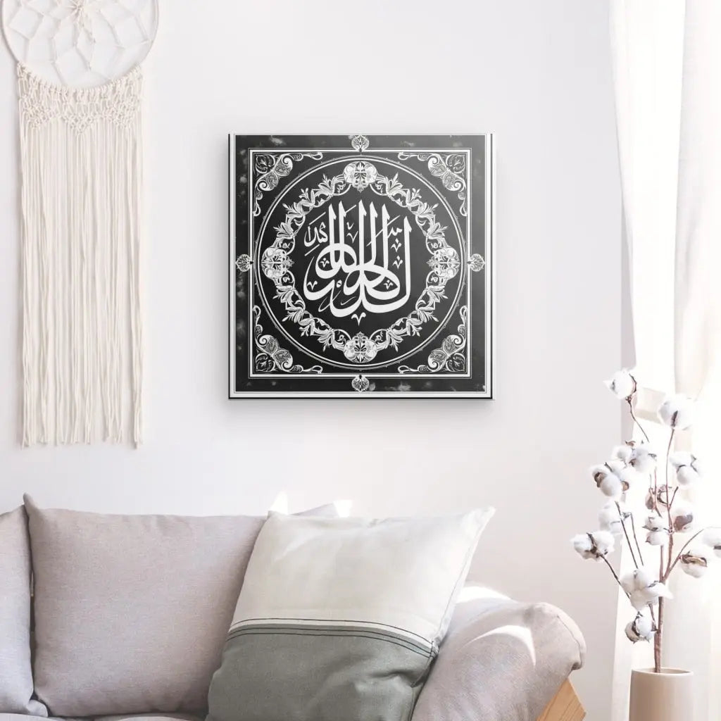 Un salon avec un canapé beige orné de coussins ; un Tableau Symbole Allah Dieu Noir et Blanc orné en noir et blanc est accroché au mur, rehaussant le design islamique de l'espace. A proximité, une décoration intérieure suspendue complète l'agencement, tandis qu'un vase avec des tiges de coton trône au premier plan.