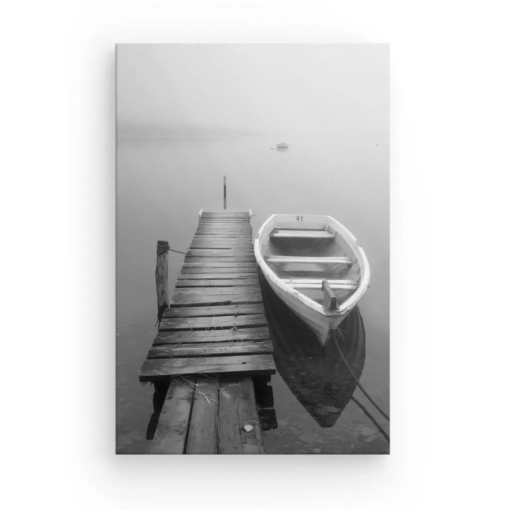 Une photo en noir et blanc capture la décoration sérénité d'un quai en bois patiné s'étendant sur des eaux calmes avec une petite barque amarrée à côté. Le brouillard obscurcit l’arrière-plan, rehaussant le paisible Tableau Ponton Barque Lac Noir et Blanc.