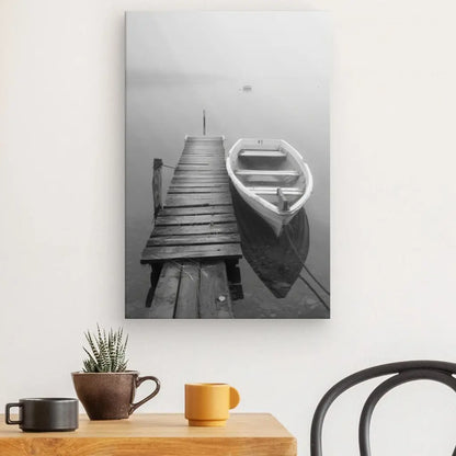 Tableau Noir et Blanc Ponton Barque Lac Noir et Blanc d'un ponton en bois avec une petite barque attachée, affiché sur un mur blanc au-dessus d'une table en bois avec une petite plante en pot et deux tasses, ajoutant de la sérénité à la décoration.