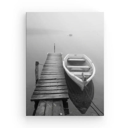 Photo en noir et blanc d’un quai en bois s’étendant sur une eau calme avec une petite barque attachée sur le côté. Un brouillard dense obscurcit l'arrière-plan, créant un Tableau Ponton Barque Lac Noir et Blanc qui ajoute à l'atmosphère tranquille et sereine, parfaite pour une décoration de sérénité.