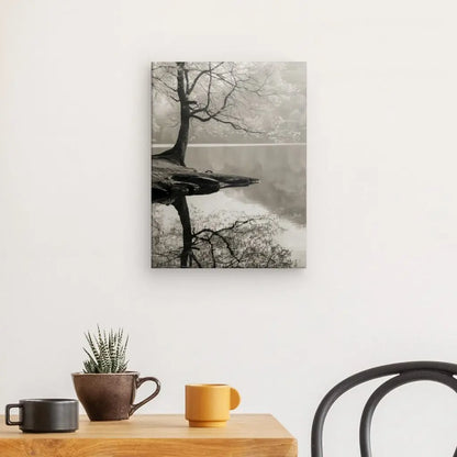 Une photographie en noir et blanc d'un arbre reflété dans un lac, ressemblant au Tableau Nature Sépia Arbre Noir et Blanc, est accrochée sur un mur blanc au-dessus d'une table en bois avec une petite plante et deux tasses. Une chaise noire est partiellement visible à droite, ajoutant à la décoration intérieure sereine.