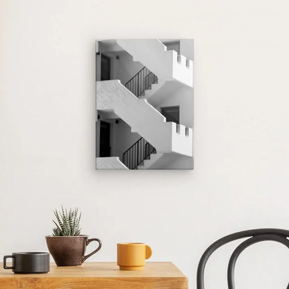 Une photo en noir et blanc d'un bâtiment avec des escaliers, intitulée "Tableau Escaliers Noir et Blanc Paysage Architectural", est accrochée sur un mur blanc au-dessus d'une table en bois ornée d'une plante, d'une tasse noire et d'une tasse jaune. Une partie d'une chaise noire est visible à droite, créant une décoration intérieure élégante.