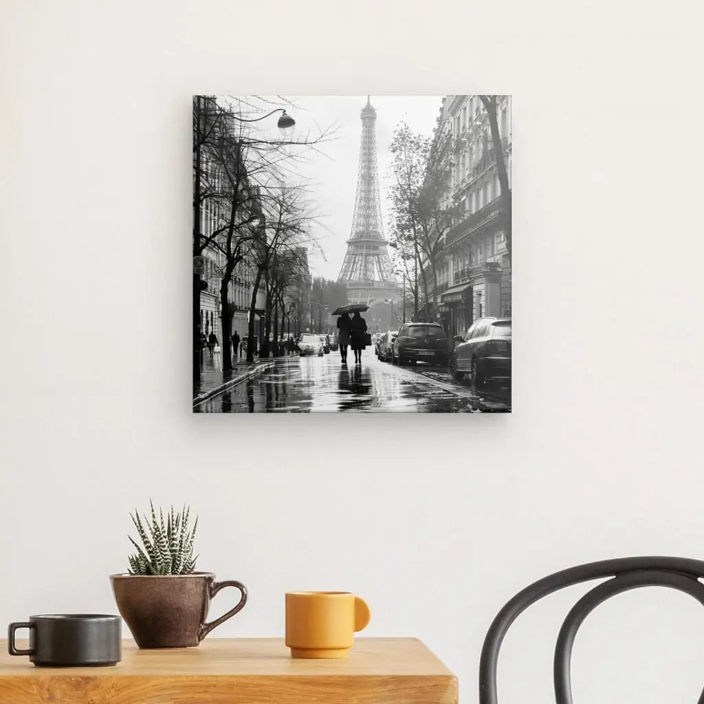 La photo en noir et blanc sur le mur représente les rues parisiennes sous la pluie, avec Tableau Paris Tour Eiffel Photo Rue Noir et Blanc en arrière-plan. A proximité, une table contient une plante en pot et des tasses. Une vue partielle d'une chaise noire est au premier plan.