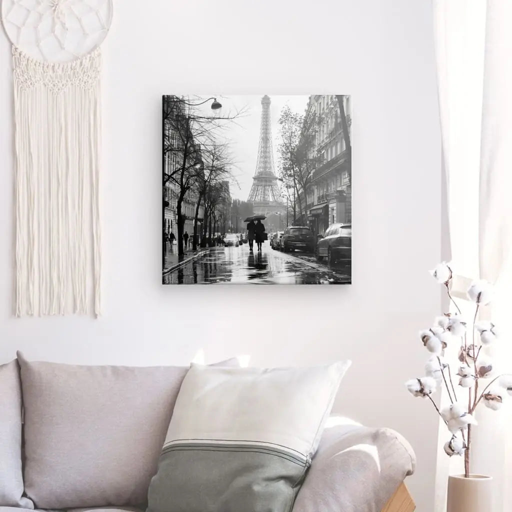 Un Tableau Paris Tour Eiffel Photo Rue Noir et Blanc orne le mur au-dessus d'un canapé gris clair avec des coussins blancs et gris. Un arrangement de cotonnier se trouve à proximité, rehaussant le tableau noir et blanc aux côtés d’une charmante tenture murale en macramé.