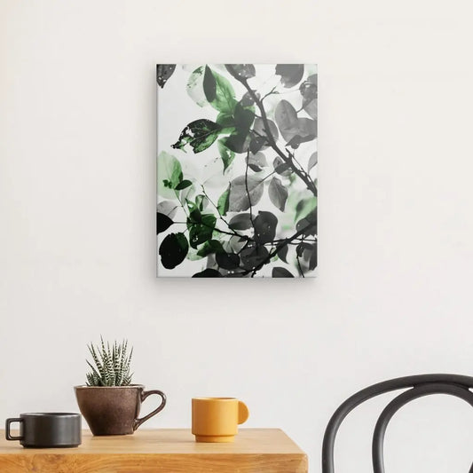 Un Tableau Nature Feuilles Noir et Blanc Touche de Vert mural avec des feuilles vertes et noires est suspendu au-dessus d'une table en bois avec une petite plante en pot et deux tasses. Une chaise noire est partiellement visible à droite, renforçant l'harmonie naturelle de la scène.